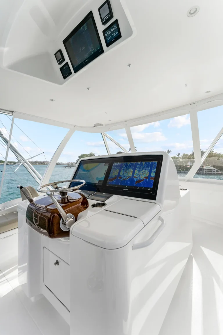 White yacht helm with wood steering wheel and multiple navigation screens, overlooking a marina.