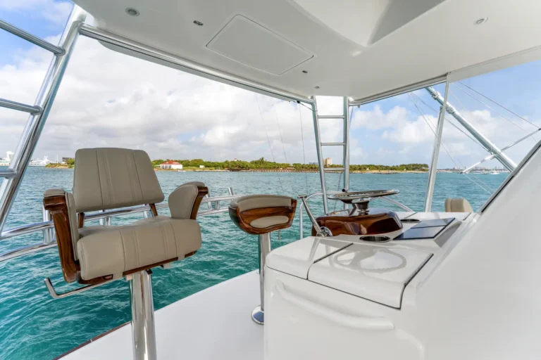 Luxury motor yacht cockpit with beige captain chairs, chrome rails, and a helm station overlooking turquoise ocean and a distant shoreline under a blue sky.