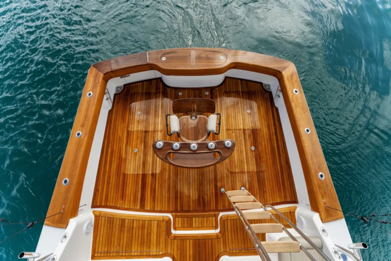 Aerial view of a polished teak boat deck with a curved seating area and steps leading down, surrounded by blue water.