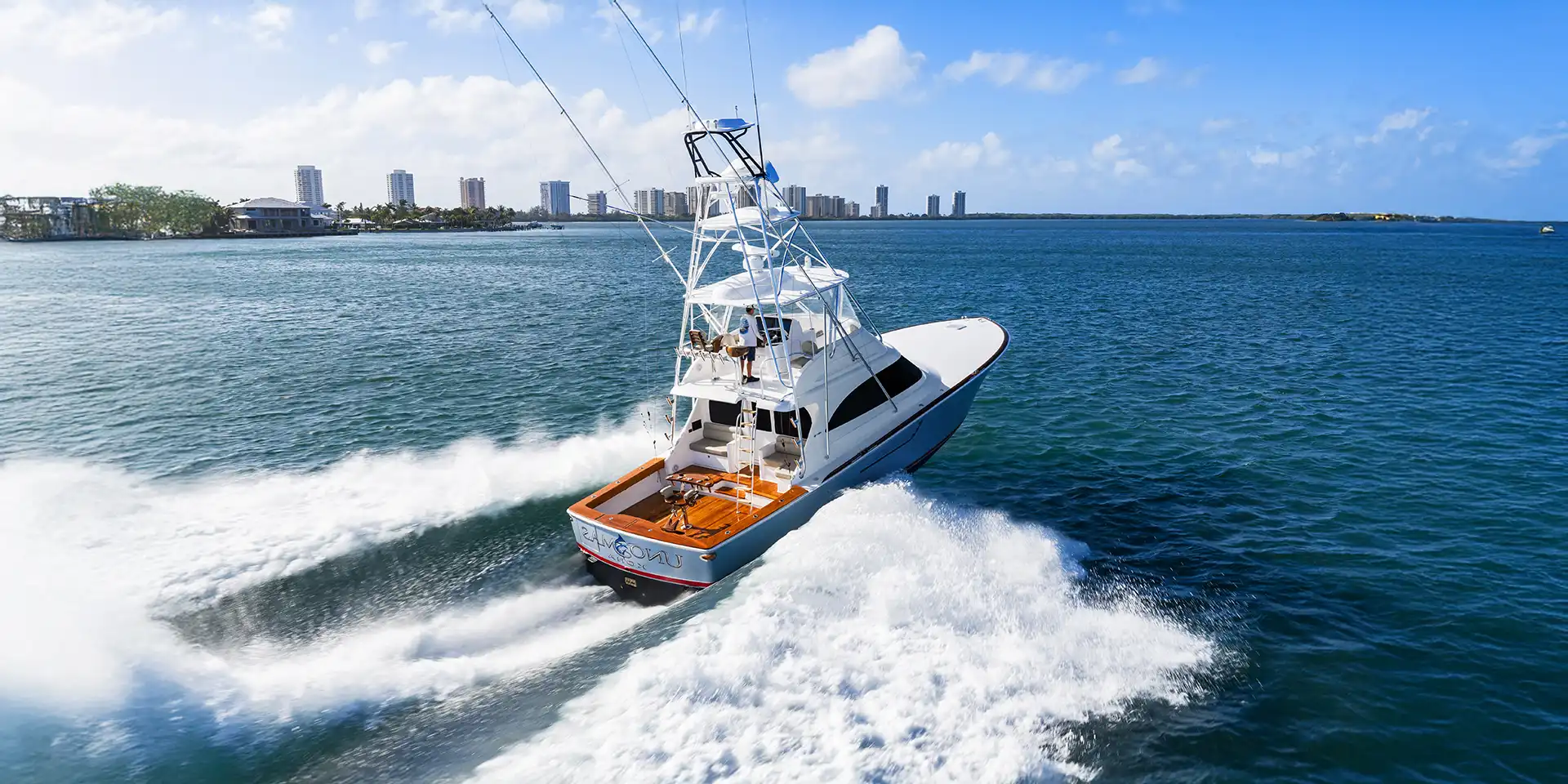 White speedboat skimming through blue water with a city skyline in the distance, under a clear sky.