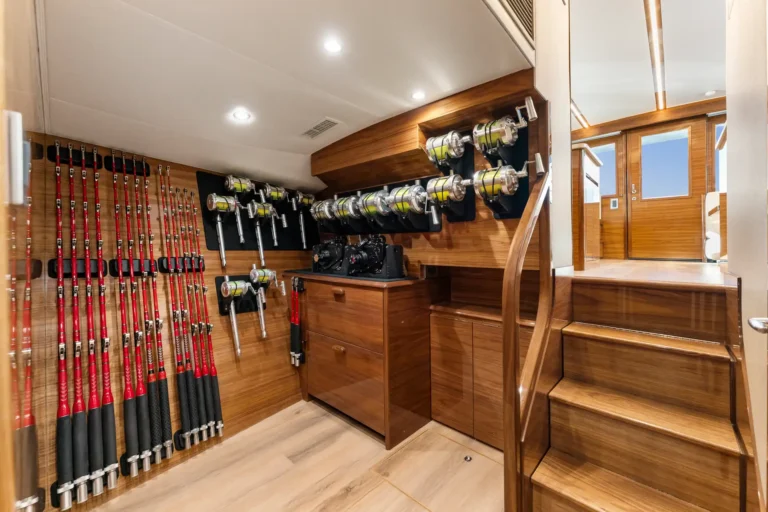 Interior of a luxury yacht galley-like room filled with fishing gear: red-and-black fishing rods along the left wall and lined-up reels on dark panels, wooden cabinets, and stairs to the right.