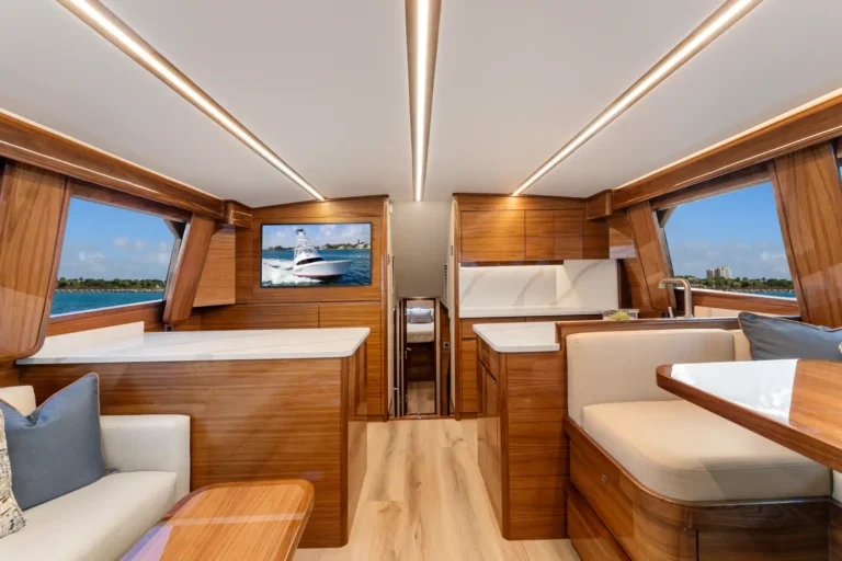 Luxurious yacht cabin with wooden panels, cream seating, and a TV mounted on the wall. Ocean view through side windows.