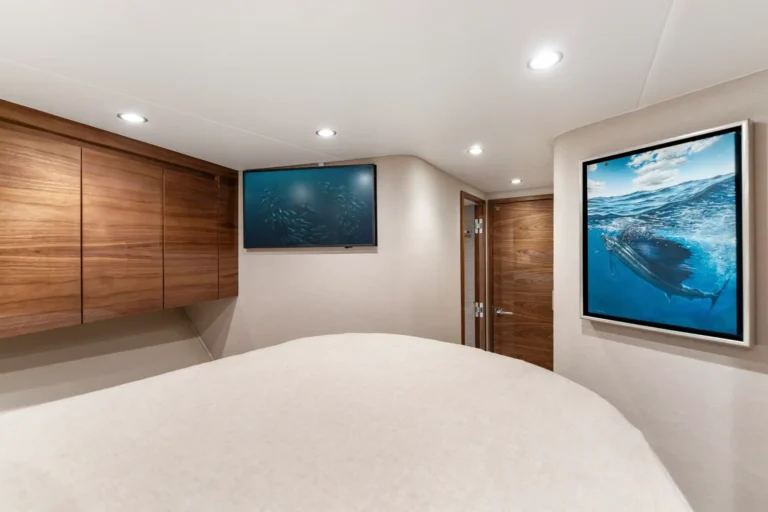 Yacht bedroom interior with wooden cabinets, recessed ceiling lights, and a bed; wall TV shows fish while a framed ocean image hangs on the right.