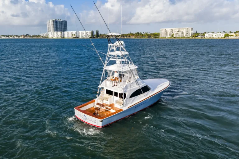 White multi-level fishing/yacht with a tall tower cruising on blue water; a city shoreline with high-rise buildings in the background.