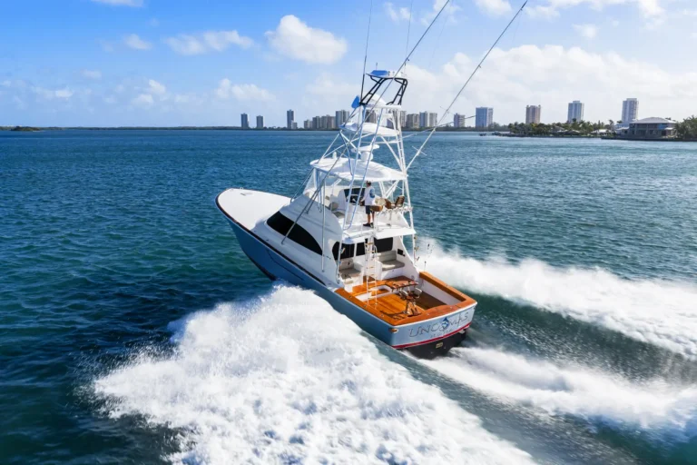 White sport fishing yacht speeds through blue water, wake spraying behind it with a city skyline in the distance.