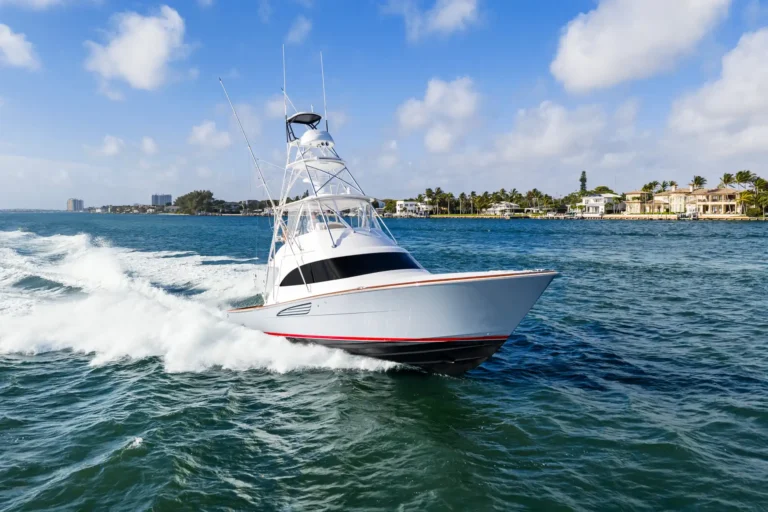 White speedboat cuts through blue ocean near a sunny shoreline with palm trees and houses in the distance.