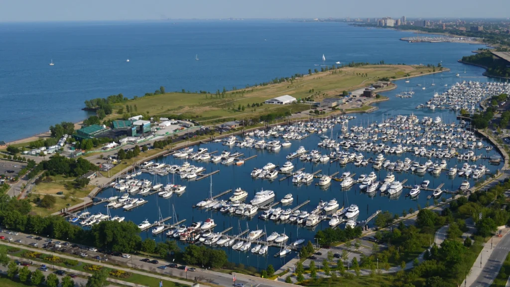Aerial view of a large marina filled with yachts and boats along a calm blue harbor with green parkland and a distant city skyline.