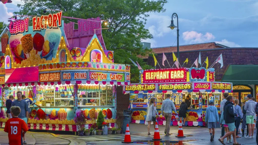 Colorful carnival stalls at dusk with bright signs for cotton candy, candy apples, and elephant ears; people strolling and cones on the wet pavement.