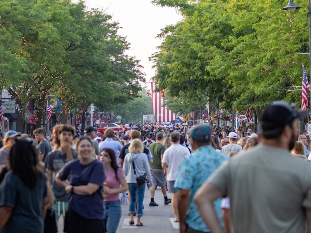 Crowded city street lined with trees and string lights, American flags, and festival banners as people walk in a parade.