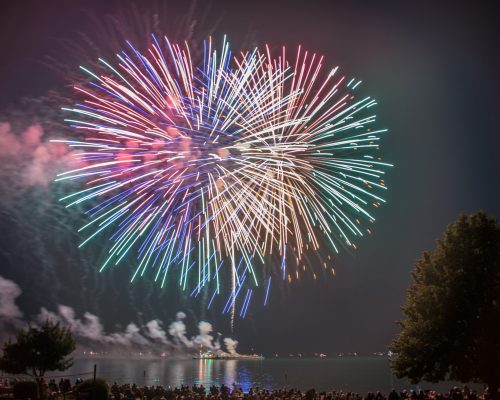 Bright multi-colored fireworks explode over a lake at night, with a crowd on the shore and trees on the right.