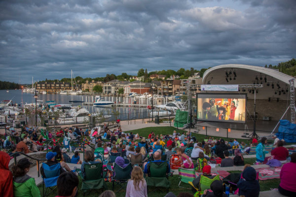 Outdoor movie night at a marina: audience seated on lawn chairs watching a movie on a stage screen beside boats under a cloudy sky.