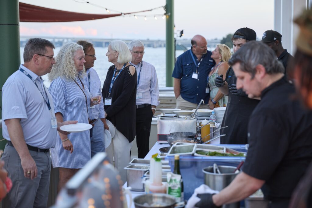 Group of adults in casual attire with name badges mingling near a seaside buffet on a dock, with a grill and plates in the foreground and water in the background.