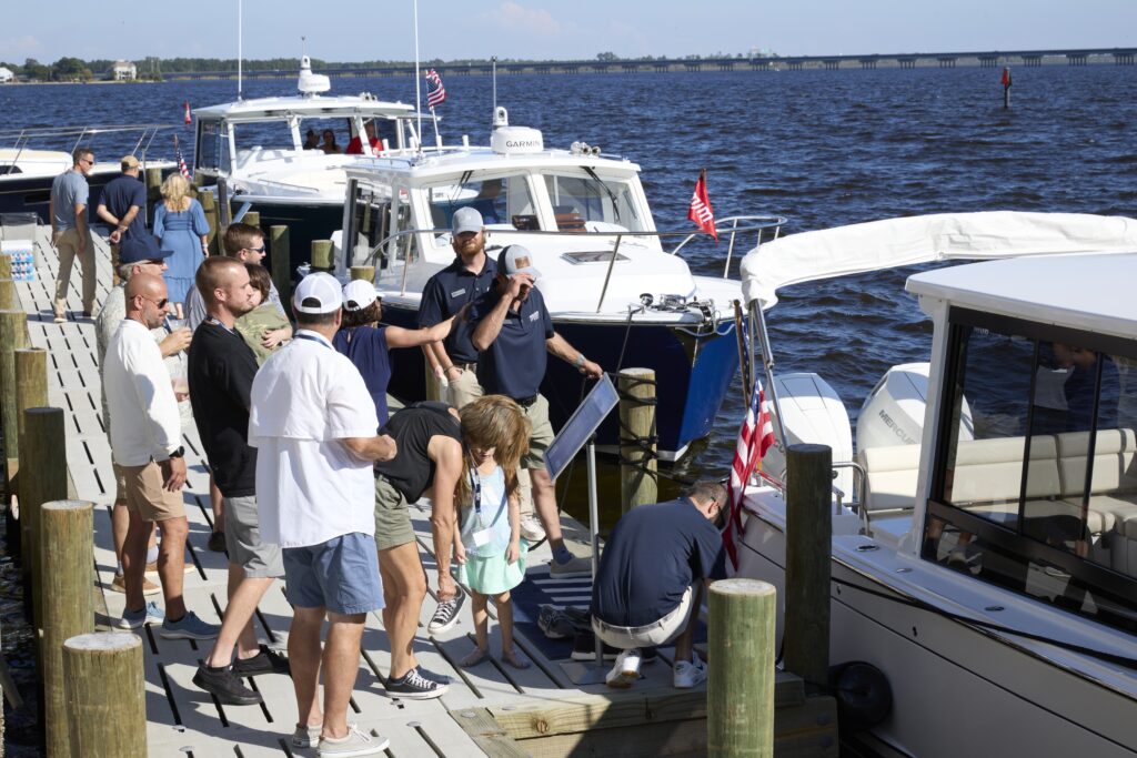 Crowd of people on a sunny marina dock beside several moored white boats with American flags nearby.