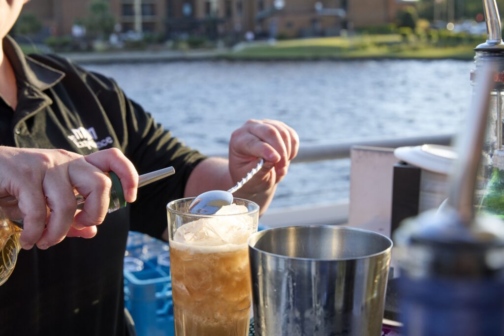 Close-up of hands pouring a drink from a shaker into a tall, ice-filled glass at an outdoor bar by the water.