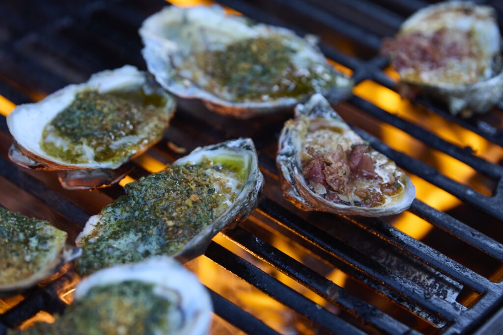 Oysters grilling on a barbecue grate topped with a green herb crust over orange flames, close-up.