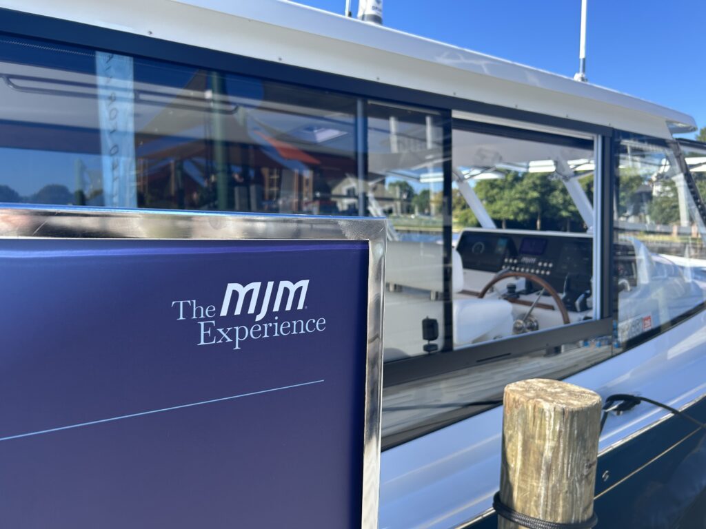 Close-up of a boat sign reading 'The MJM Experience' with water and harbor reflections in the windows behind it, and a clear blue sky above.