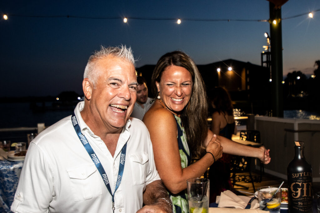 Two friends laughing and enjoying an outdoor dinner party at dusk under string lights on a waterfront deck for a festive evening.