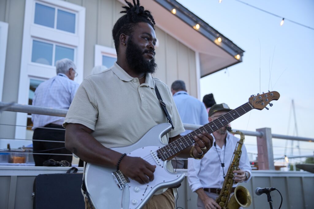 Guitarist in a beige polo shirt playing a silver electric guitar outdoors, with other band members and a saxophonist in the background under string lights.