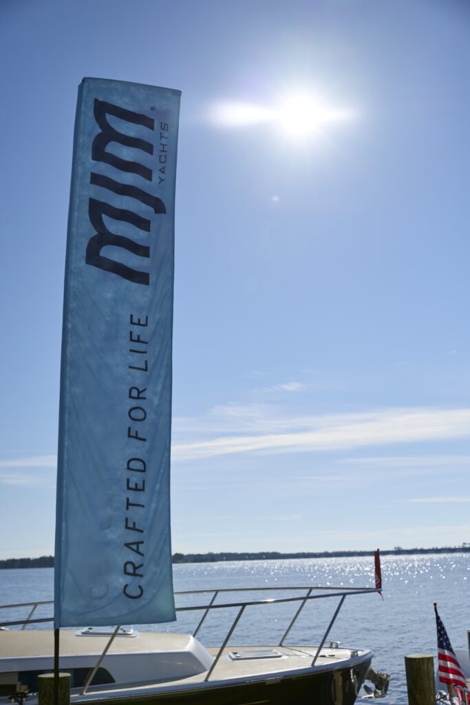 Light blue flag at a marina reading 'CRAFTED FOR YACHTS' with a sunlit body of water and boats in the background.