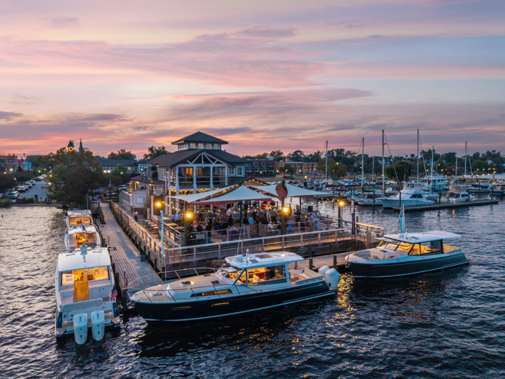 Marina at sunset with boats docked along wooden piers and a bustling waterfront patio under string lights against a pink-purple sky